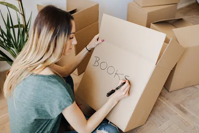 Mujer escribiendo en una caja de cartón mientras empaca libros, preparándose para el día de la mudanza en casa.